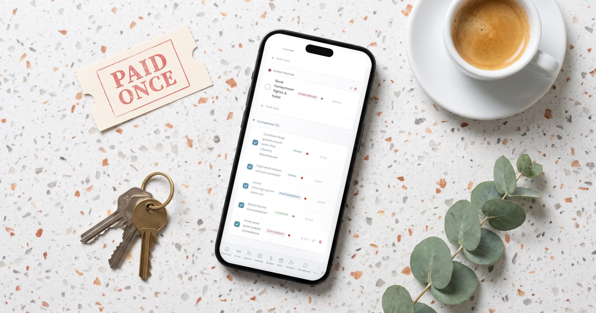 Best wedding planning apps with no subscription: a phone shows a wedding checklist app next to a paid once ticket stub, house keys and an espresso