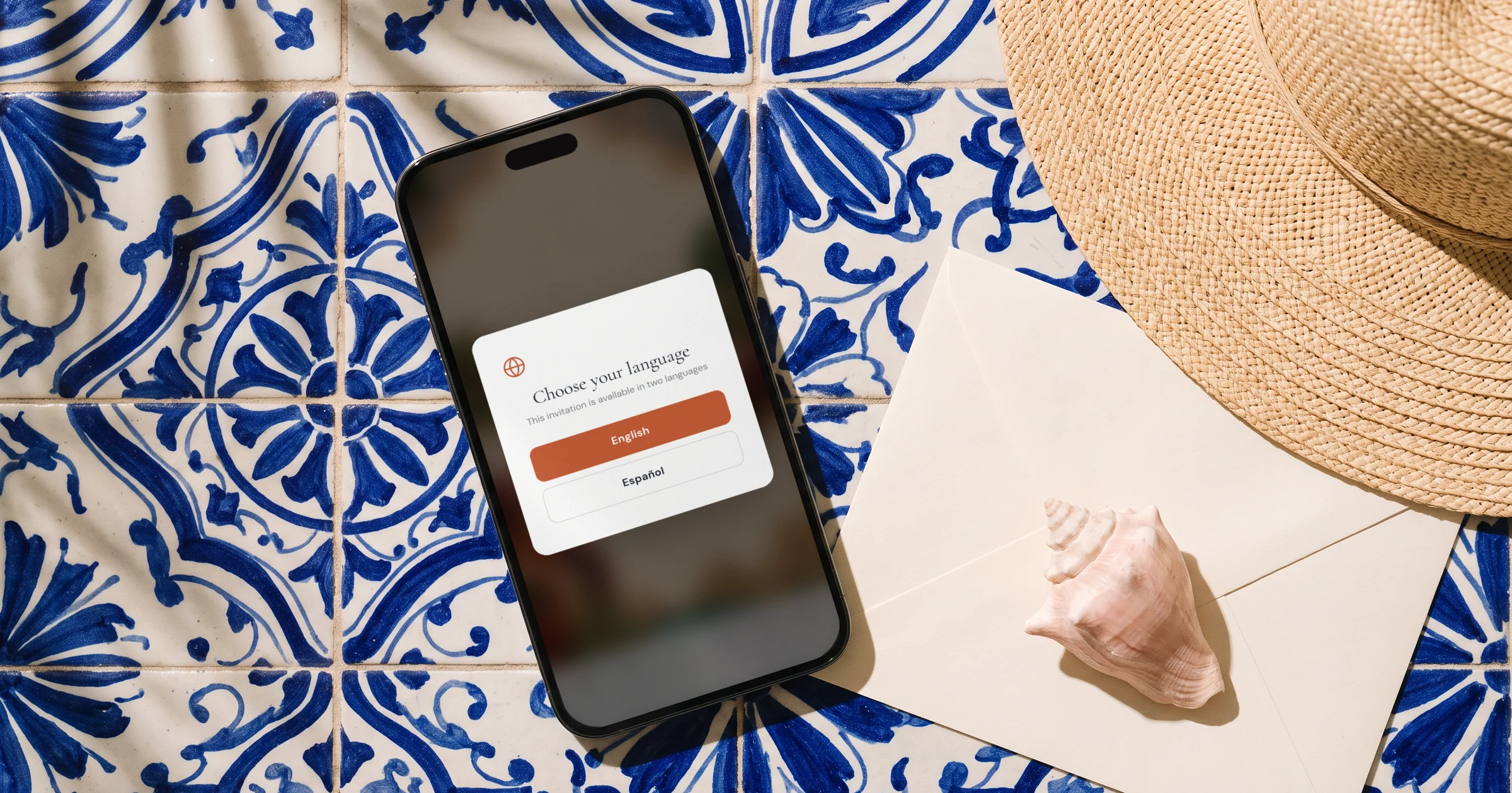 Destination wedding invitation in two languages: a phone shows the invitation asking guests to choose English or Spanish on a seaside terrace